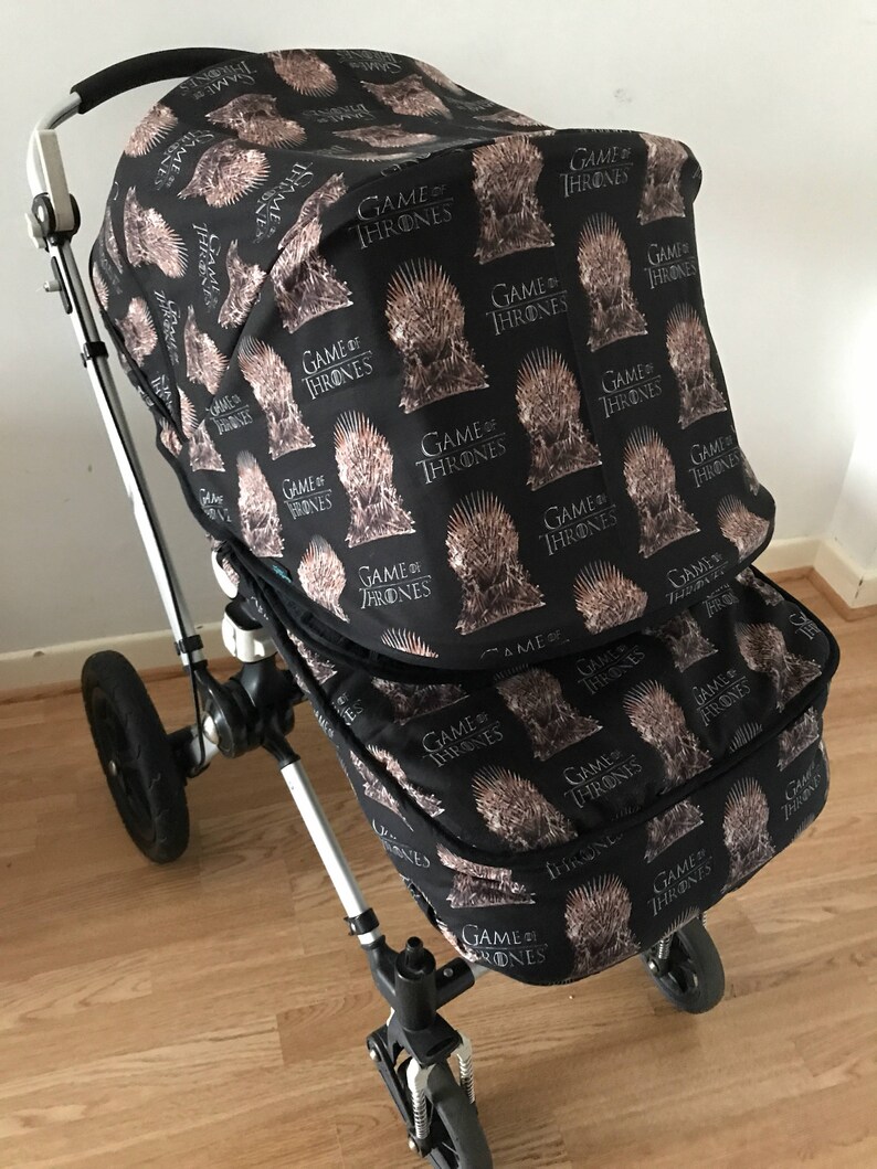May include: A black stroller with a Game of Thrones themed canopy and storage basket. The canopy and basket are covered in a repeating pattern of the Iron Throne with the words "Game of Thrones" printed on it.