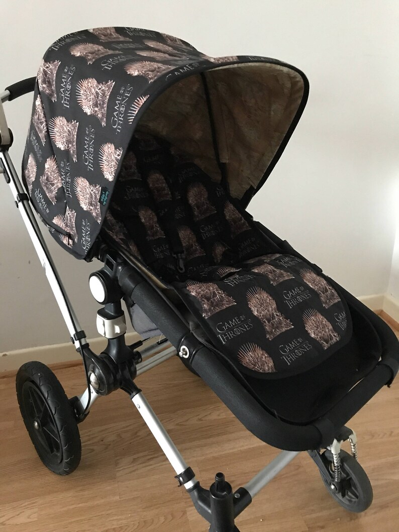 May include: A black stroller with a Game of Thrones themed canopy and seat liner. The canopy is black with a repeating pattern of the House Stark sigil, a direwolf head with a crown of thorns. The seat liner is black with a repeating pattern of the Iron Throne.