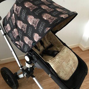 May include: A black stroller with a black and brown fabric canopy featuring a repeated pattern of the words "Game of Thrones" and a stylized image of a crown. The stroller has a black and brown fabric seat with a beige pattern.