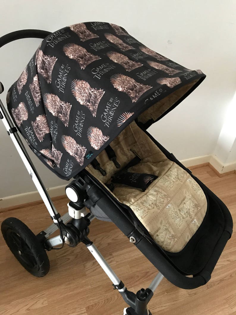 May include: A black stroller with a black and brown fabric canopy featuring a repeated pattern of the words "Game of Thrones" and a stylized image of a crown. The stroller has a black and brown fabric seat with a beige pattern.