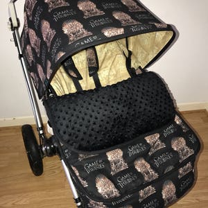 May include: A black stroller with a Game of Thrones themed canopy and seat. The canopy has a repeating pattern of the Iron Throne and the words "Game of Thrones". The seat is black with a black and white polka dot pattern. The stroller has a black basket with the same Game of Thrones pattern as the canopy.