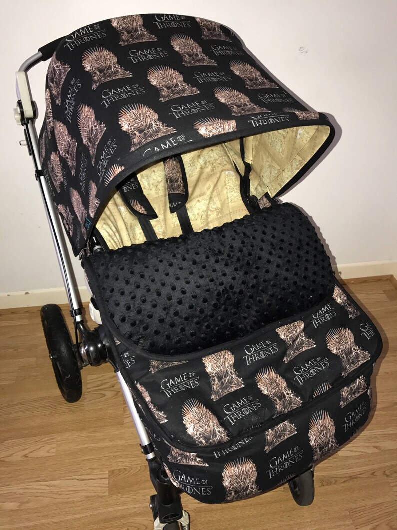 May include: A black stroller with a Game of Thrones themed canopy and seat. The canopy has a repeating pattern of the Iron Throne and the words "Game of Thrones". The seat is black with a black and white polka dot pattern. The stroller has a black basket with the same Game of Thrones pattern as the canopy.