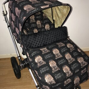 May include: A black stroller with a Game of Thrones themed fabric cover. The cover features a repeated pattern of the Iron Throne with the words "Game of Thrones" printed on it.
