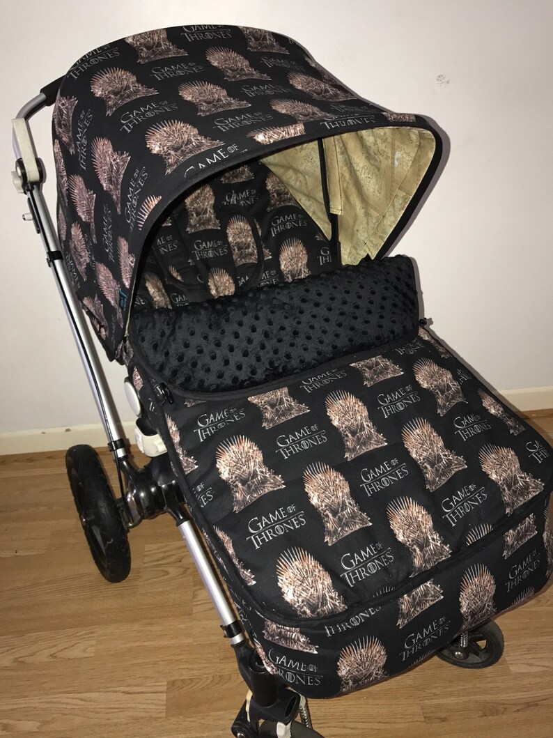 May include: A black stroller with a Game of Thrones themed fabric cover. The cover features a repeated pattern of the Iron Throne with the words "Game of Thrones" printed on it.