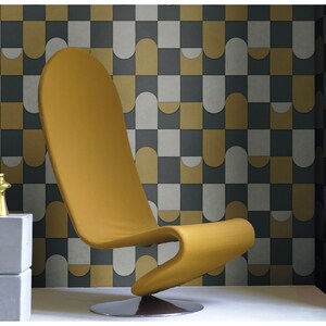 Op de afbeelding: Een mosterdgele stoel met een gebogen rugleuning en een chromen basis staat voor een muur bedekt met een geometrisch patroon van grijze, witte en gouden tegels.