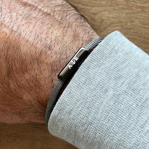 May include: A gray braided bracelet with a black metal clasp engraved with the initials "A.J.E."