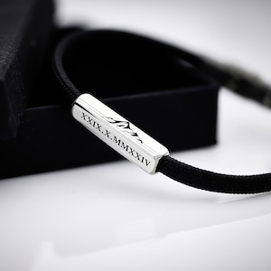 Könnte beinhalten: Ein schwarzes Kordelarmband mit einem rechteckigen Silberanhänger, der mit einer Bergsilhouette und dem römischen Datumszeichen "XIX.X.MMXIV" graviert ist.