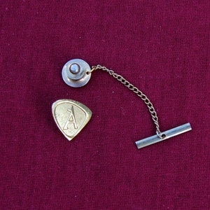 Può includere: Un fermaglio per cravatta dorato con la lettera A a forma di scudo, attaccato a una catena argentata con un fermaglio rotondo argentato.