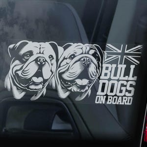 Può includere: Adesivo bianco per auto con due facce di bulldog e la scritta "BULL DOGS ON BOARD" con una bandiera Union Jack. L'adesivo è sul finestrino di un'auto, con un fanale posteriore rosso a destra.