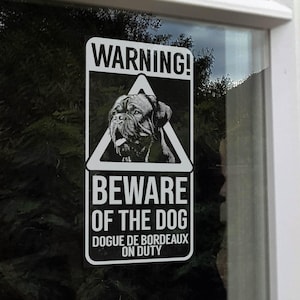 Op de afbeelding: Een rechthoekig zwart-wit waarschuwingsbord met de tekst 'WARNING!' bovenaan. Daaronder staat een driehoekige afbeelding van een hondengezicht. De tekst 'BEWARE OF THE DOG' staat onder de afbeelding, met 'DOGUE DE BORDEAUX ON DUTY' onderaan.