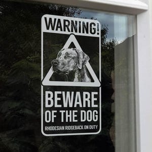 Könnte beinhalten: Ein rechteckiges schwarz-weißes Warnschild ist an einem Fenster angebracht. Das Schild zeigt "WARNUNG!" über einem dreieckigen Bild eines Hundekopfes. Darunter steht "VORSICHT VOR DEM HUND" und "RHODESIAN RIDGEBACK IM DIENST."