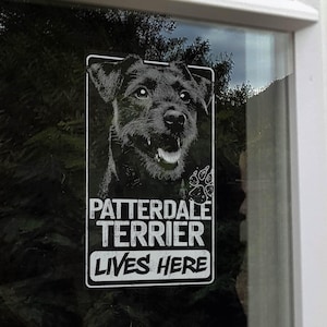 May include: A rectangular white and black sign on a window displays a Patterdale Terrier portrait and the text "PATTERDALE TERRIER LIVES HERE". The dog's image is detailed, showing fur and a happy expression, with paw prints.
