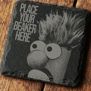 Peut inclure: Un dessous de verre en ardoise noire avec une image de Beaker des Muppets. Le dessous de verre porte l'inscription "PLACE YOUR BEAKER HERE" en blanc. Le dessous de verre est posé sur une surface en bois.