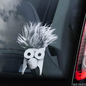 May include: White vinyl sticker of a cartoon character with spiky hair and large eyes. The character has a round nose and a wide mouth. The sticker is on a car window.