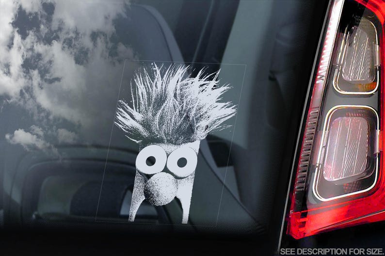 May include: White vinyl sticker of a cartoon character with spiky hair and large eyes. The character has a round nose and a wide mouth. The sticker is on a car window.