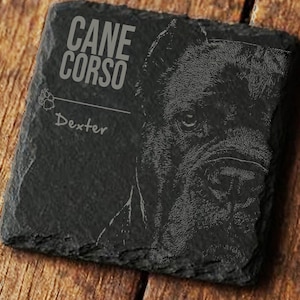 May include: A square, black slate coaster with an etched Cane Corso dog portrait. The name "Dexter" is below the dog image, with "CANE CORSO" above. The coaster is on a wooden surface, showcasing a rustic aesthetic.