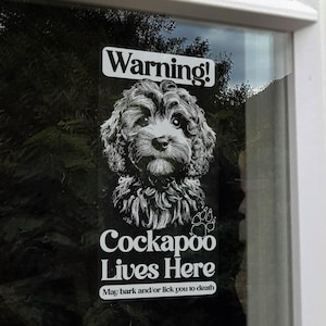 May include: A window sticker featuring a black and white Cockapoo dog illustration, with the text "Warning!" at the top. Below, it reads "Cockapoo Lives Here" and "May bark and/or lick you to death", with a paw print.