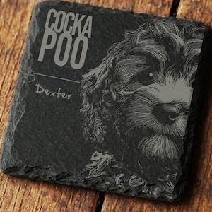 May include: A square, dark gray slate coaster with the text "COCKAPOO" and the name "Dexter" in a light gray font. A detailed illustration of a Cockapoo dog's face is on the right side. The coaster is on a wooden surface.