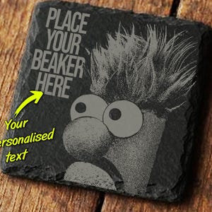 May include: A square, black slate coaster with a cartoon character design. The design features a character with large eyes and a prominent nose, with the text "PLACE YOUR BEAKER HERE". A yellow arrow points to the text, with the words "Your Personalised text".
