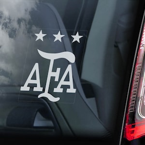 以下が含まれることがあります： 文字「AFA」と3つの星が書かれた白いビニールステッカー。ステッカーは車のリアウィンドウに貼られています。