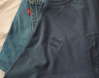 May include: A dark blue t-shirt with the text "cool dad" embroidered on the front. The t-shirt is folded and placed on top of a pair of blue jeans.