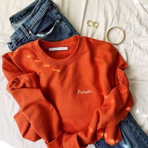 May include: A burnt orange sweatshirt with the text "Pumpkin" embroidered in white on the front. The sweatshirt is laid on a white surface with a pair of blue jeans and gold jewellery.