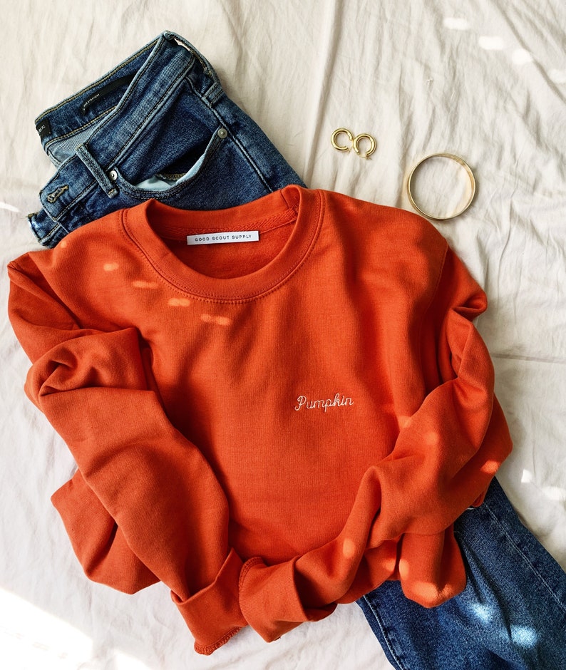 May include: A burnt orange sweatshirt with the text "Pumpkin" embroidered in white on the front. The sweatshirt is laid on a white surface with a pair of blue jeans and gold jewellery.