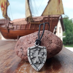 May include: A silver shield pendant with a cross design hangs from a black cord. The pendant is in focus, while a wooden ship model is blurred in the background.