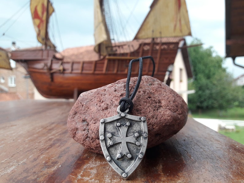 May include: A silver shield pendant with a cross design hangs from a black cord. The pendant is in focus, while a wooden ship model is blurred in the background.