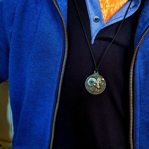 May include: A blue zippered jacket with a black shirt underneath. A silver pendant with a ram's head design hangs from a black cord.