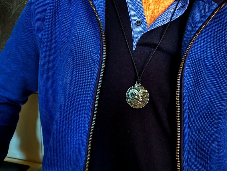 May include: A blue zippered jacket with a black shirt underneath. A silver pendant with a ram's head design hangs from a black cord.
