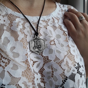 May include: A silver pendant necklace with a hexagonal charm featuring a Chinese character. The necklace is worn on a black cord and is displayed against a white lace background.