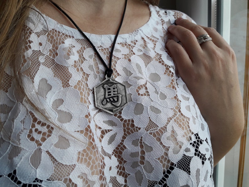 May include: A silver pendant necklace with a hexagonal charm featuring a Chinese character. The necklace is worn on a black cord and is displayed against a white lace background.