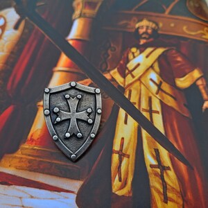 May include: A silver shield with a cross in the center, surrounded by small dots. The shield is on a background of a painting of a person in a yellow and red robe.