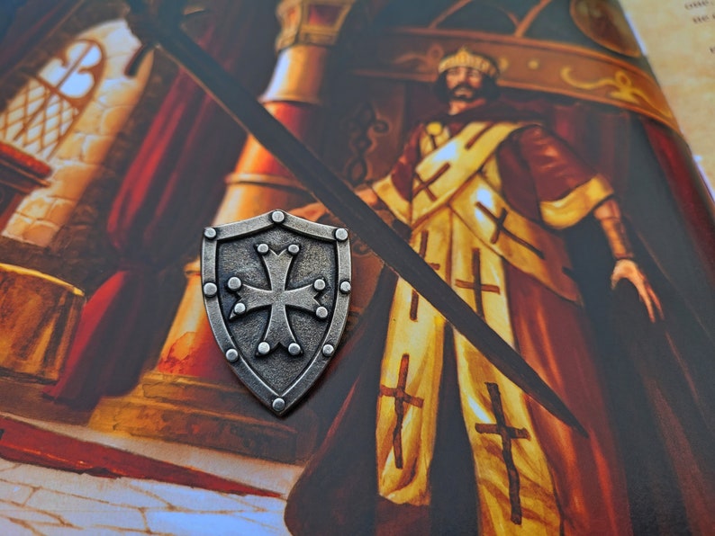 May include: A silver shield with a cross in the center, surrounded by small dots. The shield is on a background of a painting of a person in a yellow and red robe.
