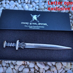 Puede incluir: Un abrecartas plateado con forma de espada en una caja negra con el texto "SWORD LETTER OPENER" e informaci&oacute;n de la tienda Etsy. El abrecartas tiene un mango detallado y una hoja puntiaguda, presentado sobre espuma negra.