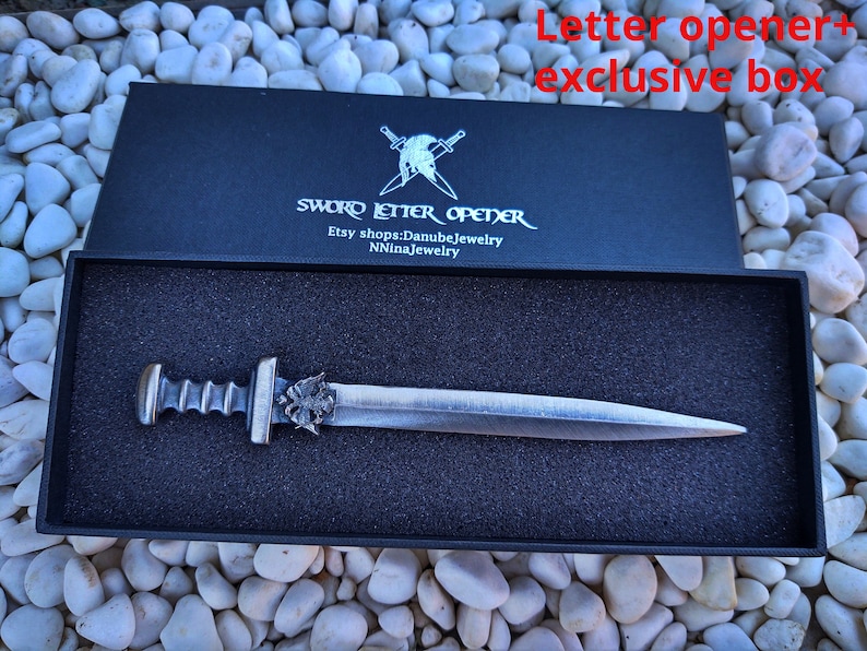 Puede incluir: Un abrecartas plateado con forma de espada en una caja negra con el texto "SWORD LETTER OPENER" e informaci&oacute;n de la tienda Etsy. El abrecartas tiene un mango detallado y una hoja puntiaguda, presentado sobre espuma negra.