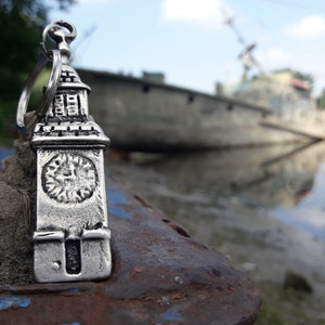 Puede incluir: Un llavero de plata con forma de torre del reloj con un diseño detallado. El llavero está sobre una superficie de metal oxidado con un fondo borroso de un barco en el agua.