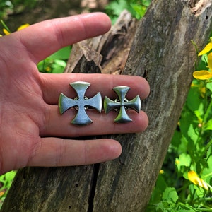 Puede incluir: Dos alfileres de metal plateado en forma de cruz, uno más grande que el otro, se sostienen en una mano contra un fondo de madera. Las cruces tienen un centro elevado y cuatro puntas.