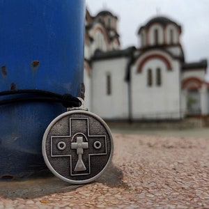 May include: A silver pendant with a cross design hangs from a black cord in front of a white church building with a blue sky in the background.