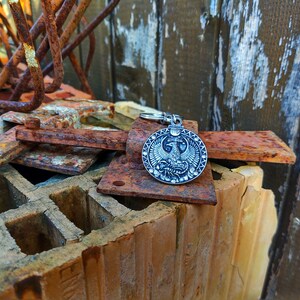 May include: A silver-toned keyring with a round pendant featuring a detailed design of a two-headed eagle with wings spread. The keyring is resting on a pile of rusty metal and brick.