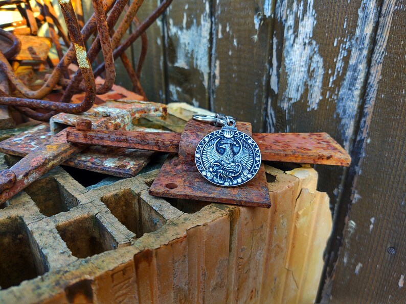 May include: A silver-toned keyring with a round pendant featuring a detailed design of a two-headed eagle with wings spread. The keyring is resting on a pile of rusty metal and brick.