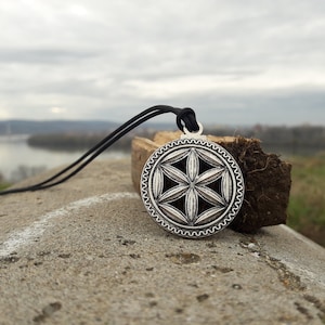 May include: A silver pendant necklace with a flower of life design. The pendant is hanging on a black cord and is resting on a piece of wood.