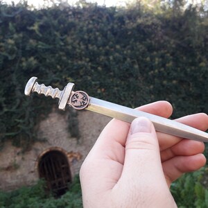 May include: A silver-colored miniature sword with a detailed hilt and a copper-colored emblem on the pommel. The sword is held in a hand against a blurred background of green foliage and a stone wall.