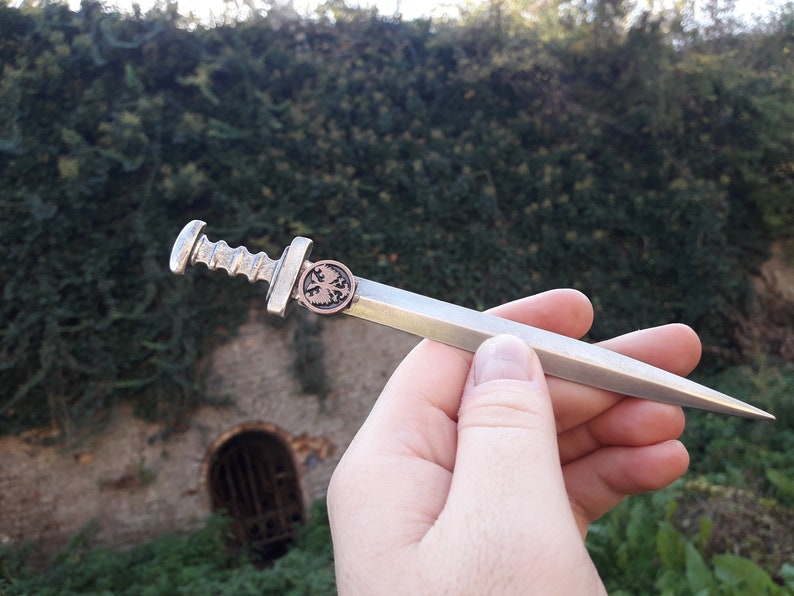 May include: A silver-colored miniature sword with a detailed hilt and a copper-colored emblem on the pommel. The sword is held in a hand against a blurred background of green foliage and a stone wall.