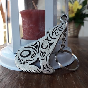 Peut inclure: Un porte-clés en argent en forme de loup avec un motif tribal détaillé. Le loup est tourné vers le haut, la gueule ouverte, et le porte-clés est attaché à un anneau en argent.