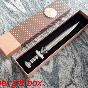 Puede incluir: Un abrecartas plateado en una caja de regalo decorativa. El abrecartas tiene un mango detallado y una hoja. La caja de regalo es marr&oacute;n con un patr&oacute;n de puntos blancos y una tapa marr&oacute;n claro.