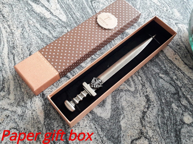 Puede incluir: Un abrecartas plateado en una caja de regalo decorativa. El abrecartas tiene un mango detallado y una hoja. La caja de regalo es marr&oacute;n con un patr&oacute;n de puntos blancos y una tapa marr&oacute;n claro.