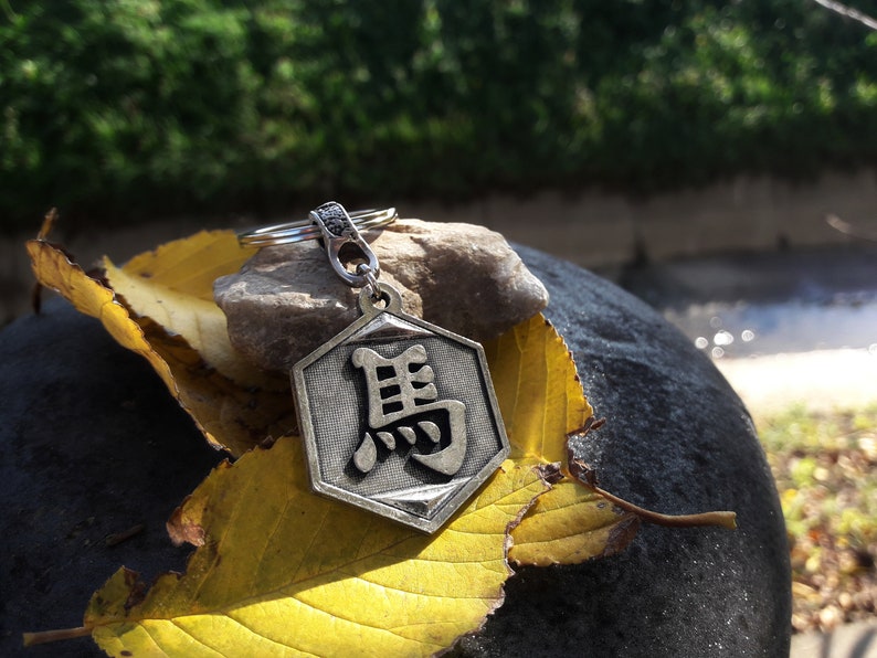 May include: A silver-colored keychain with a hexagonal charm featuring a Chinese character. The keychain is resting on a rock and a yellow leaf.
