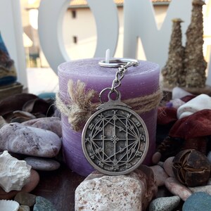 May include: A silver charm with a geometric design hangs from a silver key ring. The charm is in front of a purple candle with a brown twine bow.